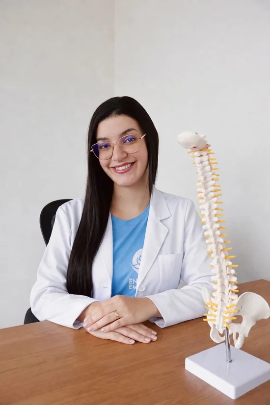 Natália Gregorio de jaleco, sentada à mesa ao lado de um modelo de coluna vertebral